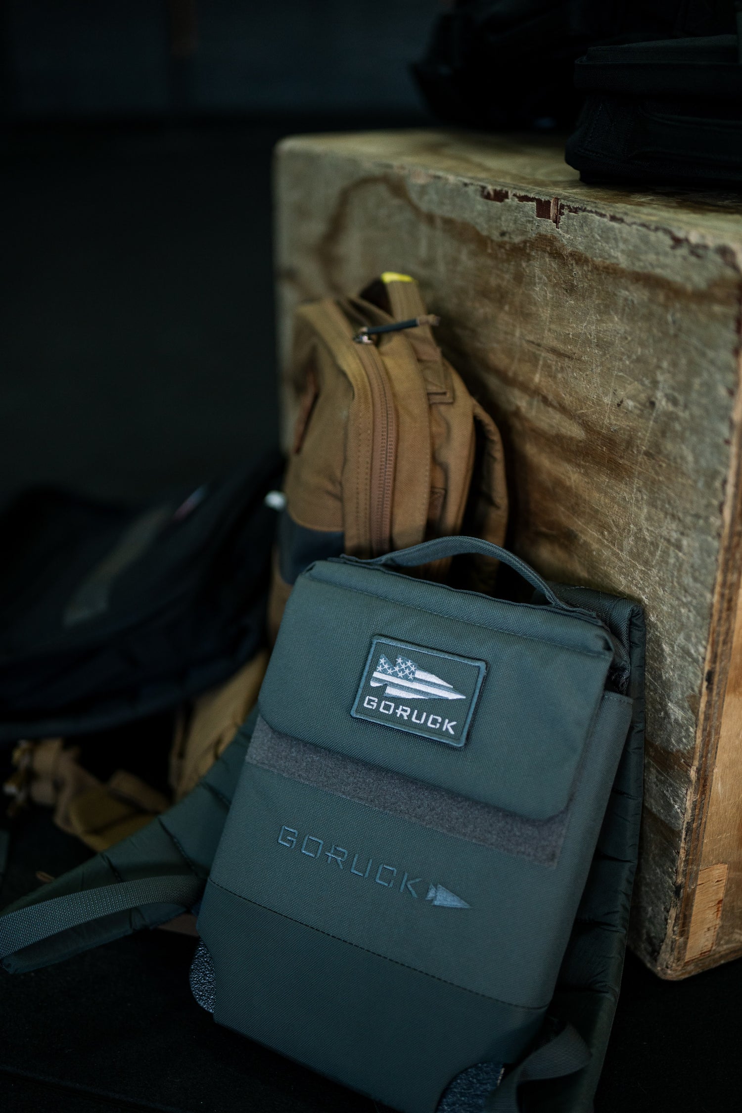 An RPC 3.0 + 10lb Ruck Sandplate sits beside a brown bag against a worn wooden box in a gym setting.