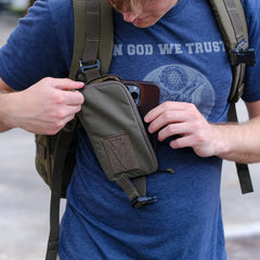 Person in navy blue 'In God We Trust' t-shirt placing phone in olive green tactical chest pouch attached to backpack strap