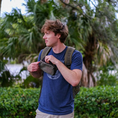 Young man outdoors wearing a tactical backpack and opening a small zippered pouch against a backdrop of green trees