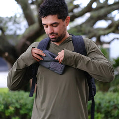 Man outdoors adjusting gray chest pouch on olive green shirt with black rucking backpack and tree background