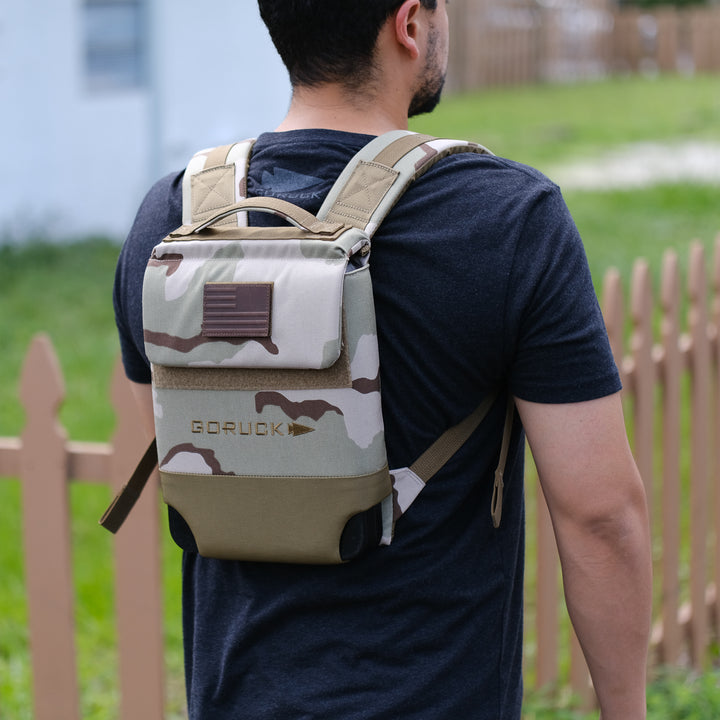 A man stands outside near a wooden fence, wearing a small camo-patterned Ruck Plate Carrier 3.0 made from rugged ballistic nylon.