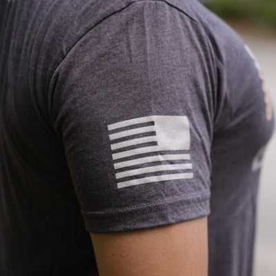 A white American flag graphic accents the sleeve of the GORUCK Logo DCU Camo Tee - Tri-Blend, a dark gray Special Forces-inspired T-shirt.
