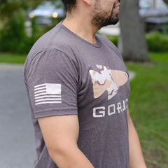 Man outdoors wearing the GORUCK Logo DCU Camo Tee, a gray tri-blend T-shirt with camo fish graphic, faded flag on the sleeve, and subtle spearhead logo nodding to Special Forces.