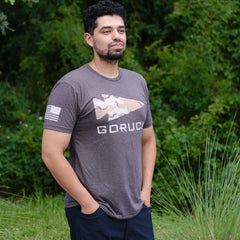 Man with short curly hair and beard stands outdoors, hands in pockets, wearing a GORUCK Logo DCU Camo Tee - Tri-Blend and shorts.