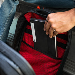 A person places a black notebook into a Bullet Ruck x Carryology - GRXC2 Samurai 2.0 backpack, showcasing the limited-edition model’s red patterned interior and unique style.