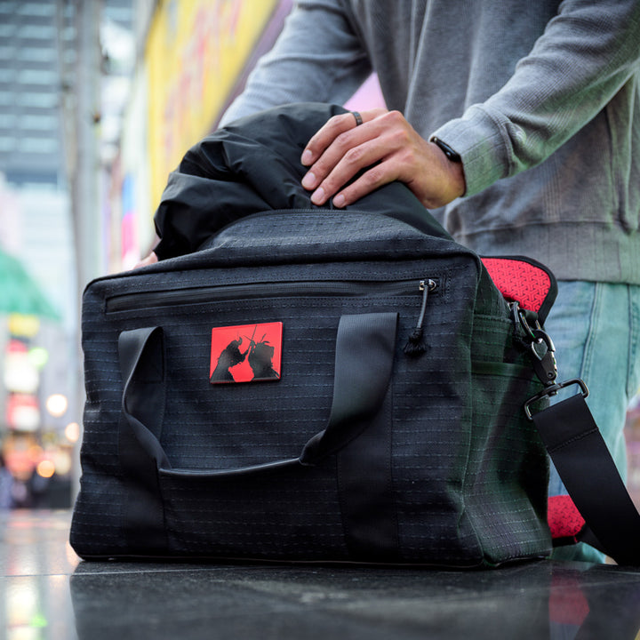 A person places a jacket into the Kit Bag x Carryology - GRXC2 Samurai 2.0, a black duffel with a red patch made from custom technical fabric, while standing on a city street.