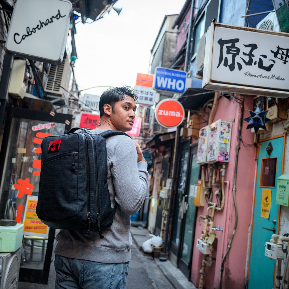 A man with a backpack, clad in GR2 x Carryology - GRXC2 Samurai 2.0 denim, walks through a narrow, colorful alley lined with various shop signs.