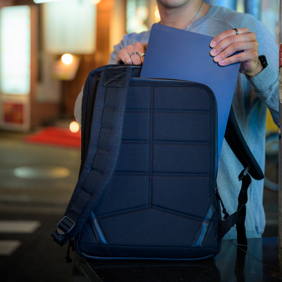 A person places a laptop into a dark blue GR2 x Carryology - GRXC2 Samurai 2.0 backpack at night in an urban setting, wearing JPXDP denim from the exclusive Japan Blue collaboration.