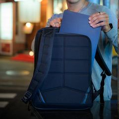 Man placing blue laptop into the back compartment of a black GORUCK backpack in urban setting