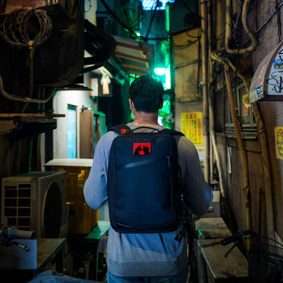 A person with a backpack, clad in the GR2 x Carryology - GRXC2 Samurai 2.0, walks through a narrow, dimly lit urban alley at night.