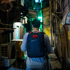 Man wearing GORUCK backpack walking through narrow, dimly lit urban alley with neon lights