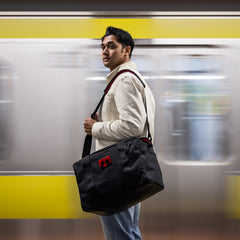 Man standing in subway station with black GORUCK duffel bag and beige jacket against yellow train