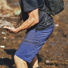 A person wearing Men’s Challenge Shorts - Lightweight ToughDry® in blue and a black shirt lifts a muddy log outdoors, with dirt on their clothes and skin.