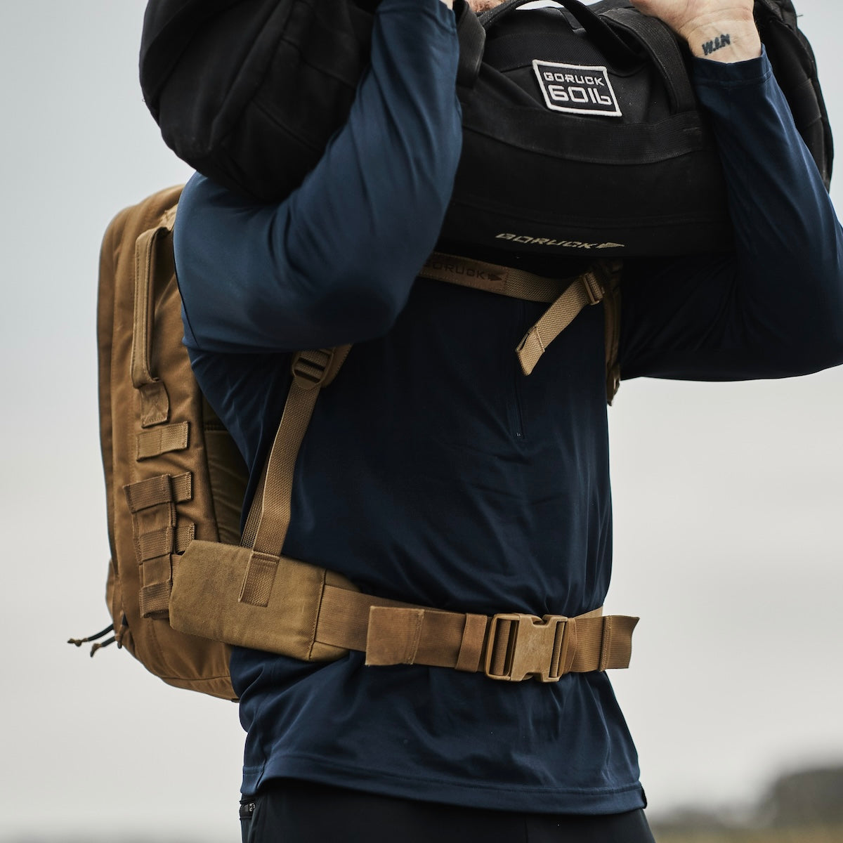 Person wearing a tan backpack and carrying a black sandbag over their shoulders outdoors.