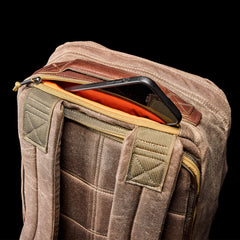 The GR1 USA - Heritage Waxed Canvas rucksack, featuring an orange lining, is shown unzipped with a black tablet inside against a black background.