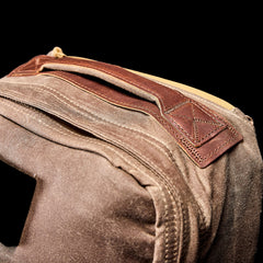 Close-up of the GR1 USA - Heritage Waxed Canvas rucksack, made from waxed duck canvas with a leather handle and yellow zipper, shown against a black background.