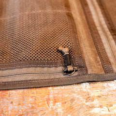 Close-up of the Wire Dopp - Waxed Canvas by GORUCK, featuring a brown mesh fabric with a zipper and toggle fastening on a wooden surface, highlighting its intricate stitching. Perfect for travel essentials, the textured design ensures optimal organization while providing durability similar to waxed canvas.