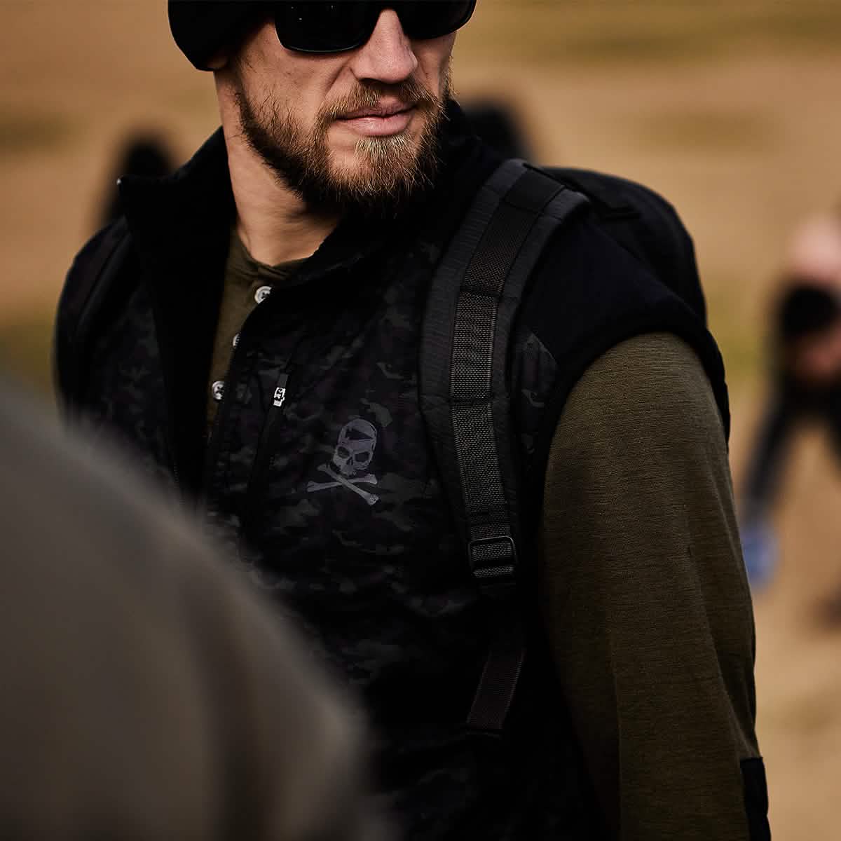 A bearded man in sunglasses and a dark beanie strides outdoors, wearing a camouflage-patterned vest adorned with a skull emblem and carrying his reliable GORUCK Bullet Ruck Double Compartment - Dyneema. In the blurred, sandy backdrop, people move around him, unaware of the rugged durability and lifetime guarantee of his gear.