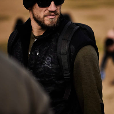 A bearded man in sunglasses and a dark beanie strides outdoors, wearing a camouflage-patterned vest adorned with a skull emblem and carrying his reliable GORUCK Bullet Ruck Double Compartment - Dyneema. In the blurred, sandy backdrop, people move around him, unaware of the rugged durability and lifetime guarantee of his gear.