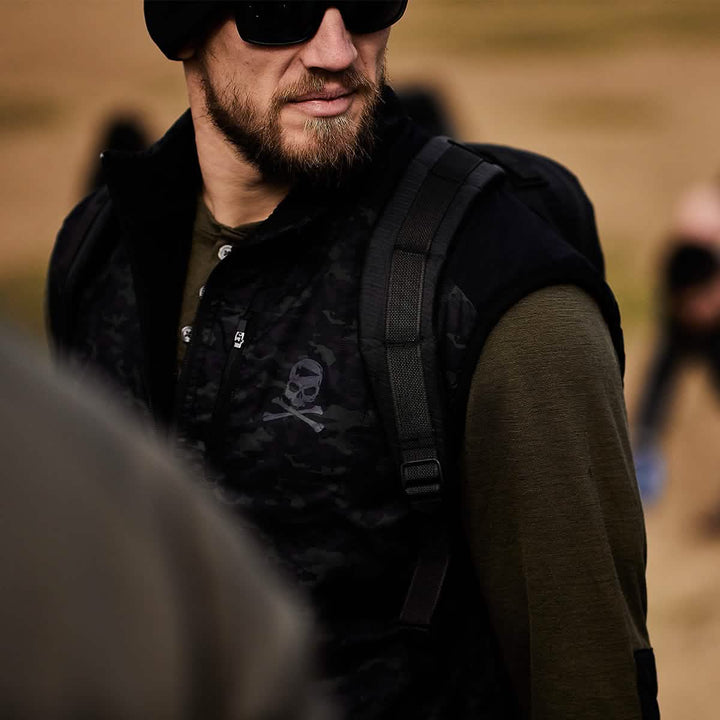 A bearded man in sunglasses and a dark beanie strides outdoors, wearing a camouflage-patterned vest adorned with a skull emblem and carrying his reliable GORUCK Bullet Ruck Double Compartment - Dyneema. In the blurred, sandy backdrop, people move around him, unaware of the rugged durability and lifetime guarantee of his gear.