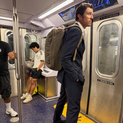 A man in a suit stands in a subway carrying the GR1 USA - Heritage Waxed Canvas rucksack, while another person sits nearby, looking at their phone.