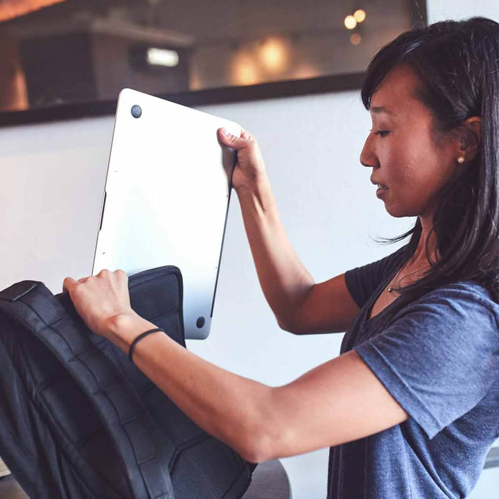 A woman in a blue shirt carefully places her silver laptop into the bombproof laptop compartment of the GR0 - Cordura backpack by GORUCK. She appears focused, preparing to head out, set indoors with a blurred background accentuating her minimalist EDC approach.
