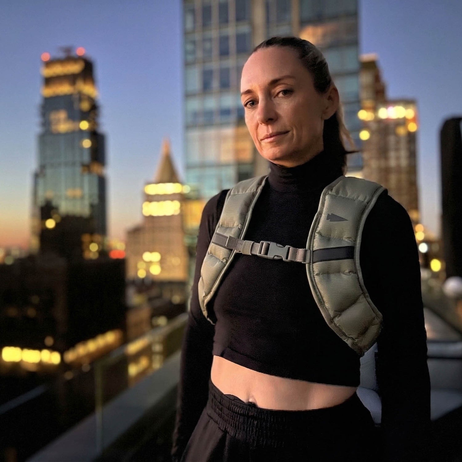 A woman in a crop top and the Spy Ruck | Women's Weighted Vest stands on a rooftop at dusk, illuminated by the city's skyscrapers.