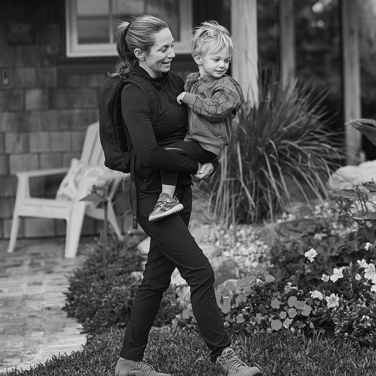 A woman carrying a young child walks past a garden, both smiling in an outdoor setting.