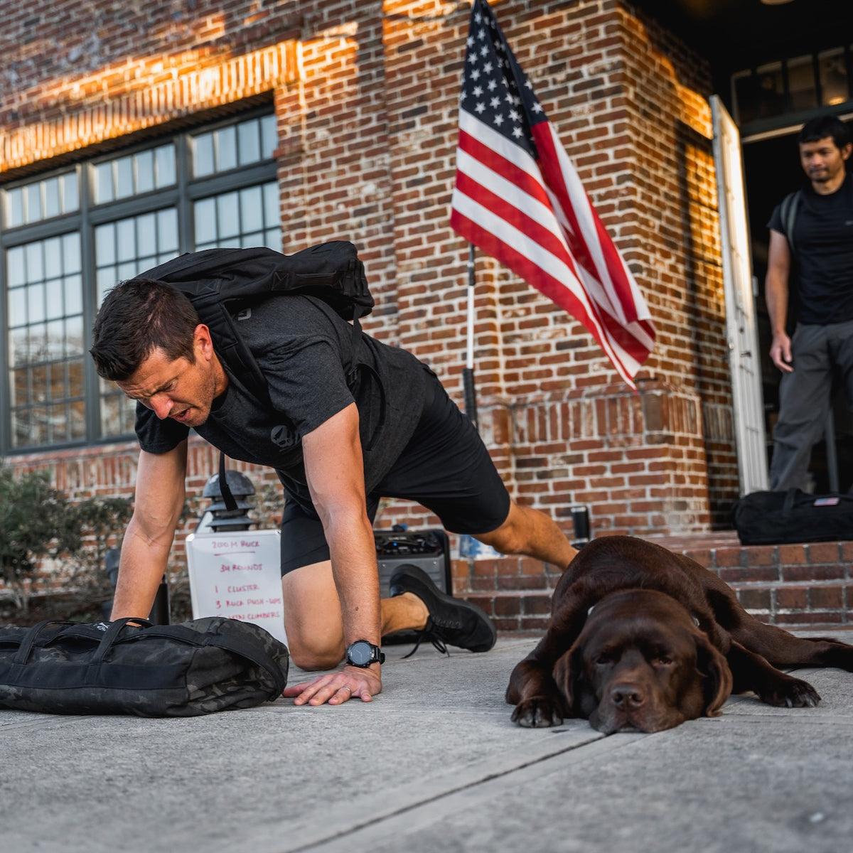 Wearing the GORUCK Tribe Tee - Tri-BlendX, a man does push-ups on the sidewalk next to a resting brown dog, with the U.S. flag and another man in the background—highlighting the true sense of community at every GORUCK Challenge.