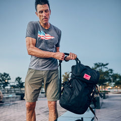 A man in a grey graphic t-shirt and 2-way stretch fabric green shorts adjusting the straps of a black GORUCK backpack for warm weather rucking.
