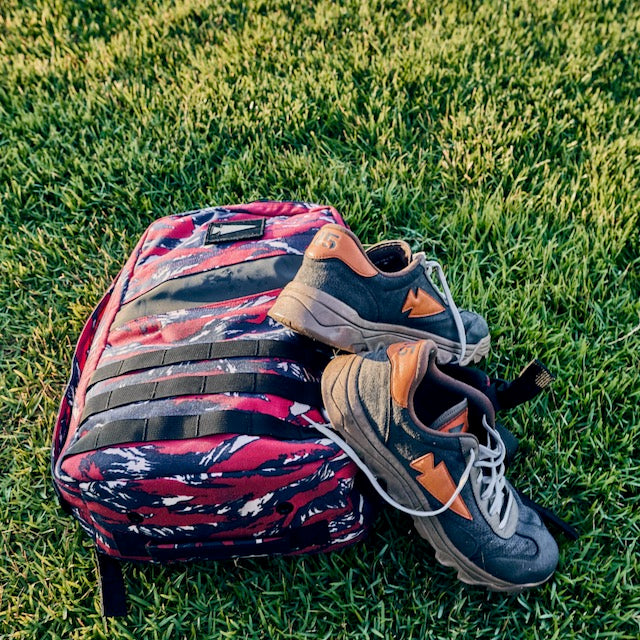 Resting on green grass are the Mackall 45 Fog—gray sneakers with orange accents and a heritage suede upper—and a red camouflage backpack inspired by the Special Forces rucking chassis.