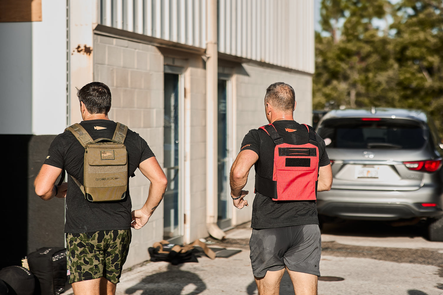 Two men wearing RPC 3.0 + 10lb Ruck Sandplate jog outside by a building and silver SUV on a sunny day, adding weighted training to their routine.