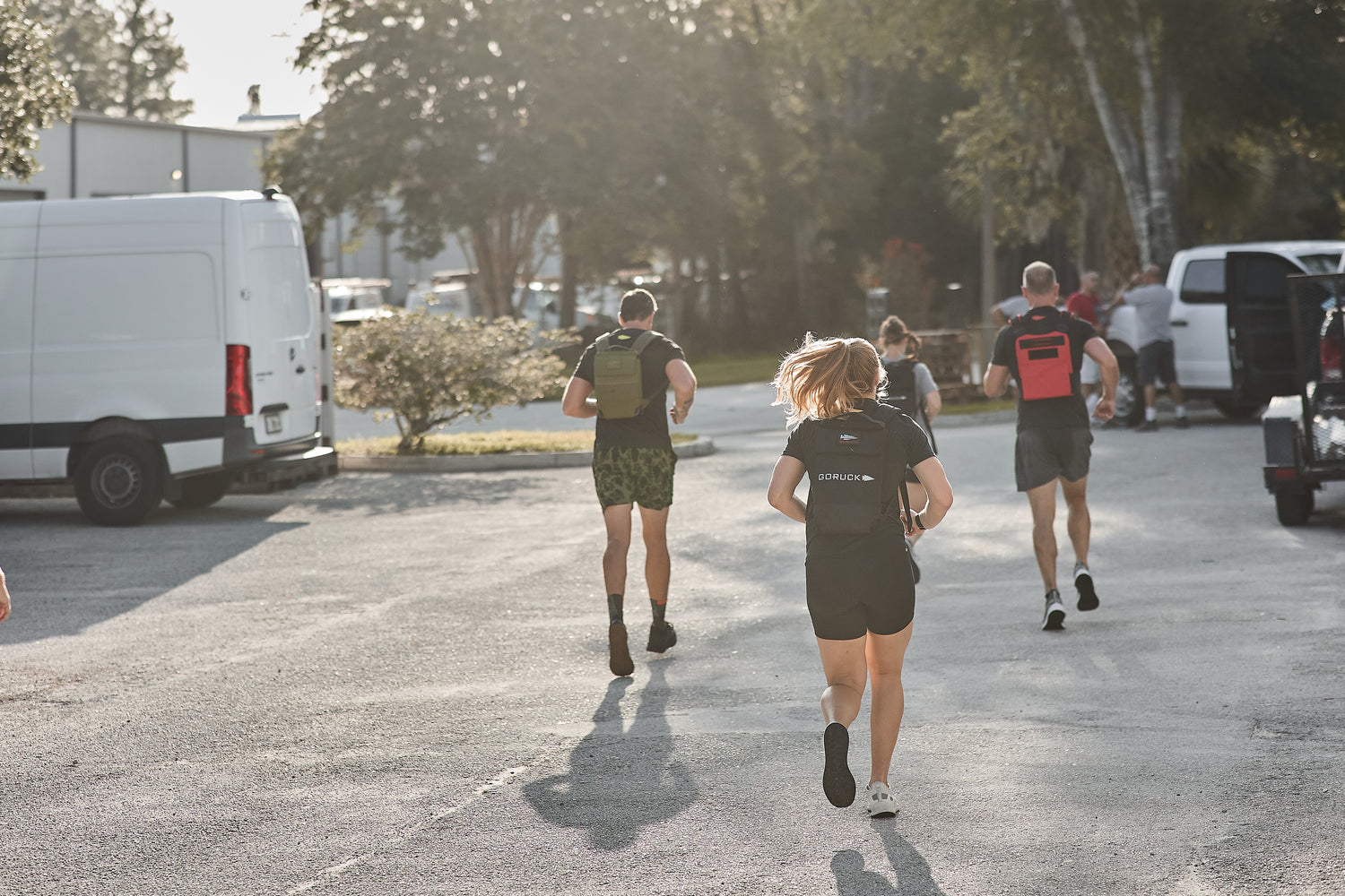 People jogging outdoors on a sunny day, wearing the RPC 3.0 + 10lb Ruck Sandplate for weighted training, with cars and trees in the background.