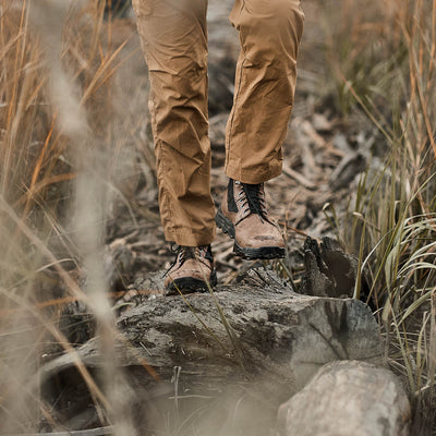 Someone wearing the MACV-2 - Mid Top in Chestnut + Black, featuring an aggressive triple compound outsole, pairs them with khaki pants while strolling along a wooded path bordered by tall grass.