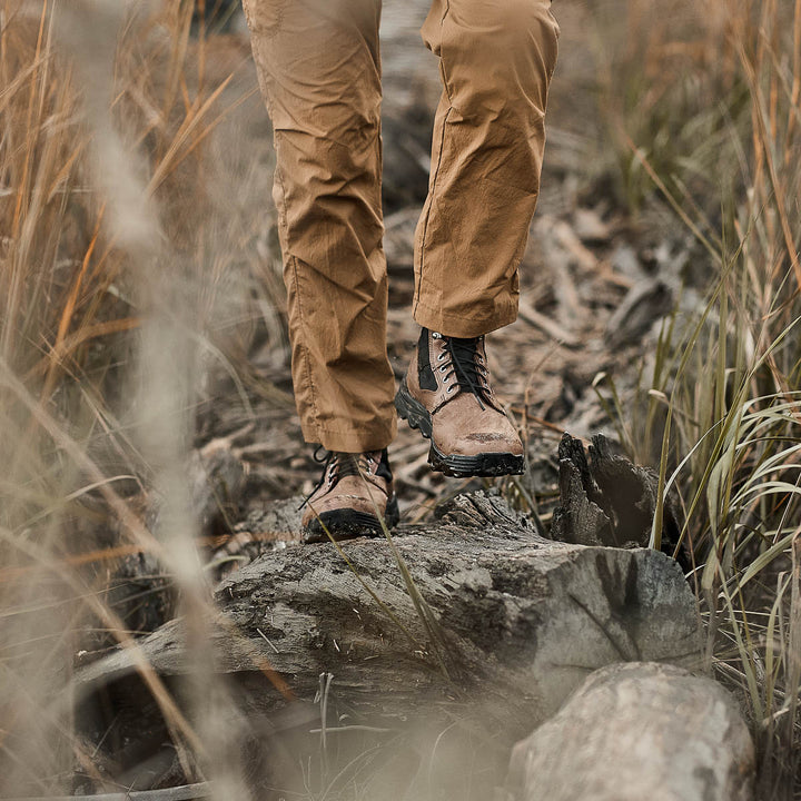 Someone wearing the MACV-2 - Mid Top in Chestnut + Black, featuring an aggressive triple compound outsole, pairs them with khaki pants while strolling along a wooded path bordered by tall grass.