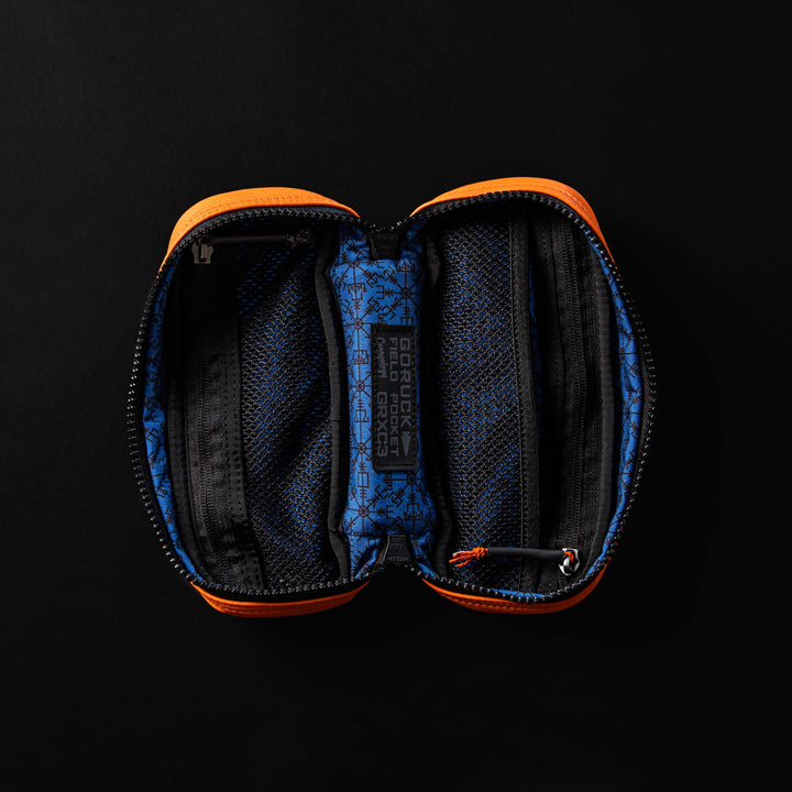 The Echo Field Pocket x Carryology - Berserker Viking by GORUCK is an open zippered pouch adorned with orange edges and a blue interior showcasing black Viking-inspired patterns. Inside, it boasts two main mesh compartments alongside a central fabric label with inscriptions. Set against a striking black background, it's designed to resonate with the spirit of Norsemen.