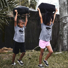 Two children in gray GORUCK t-shirts lifting black rucking bags outdoors during training