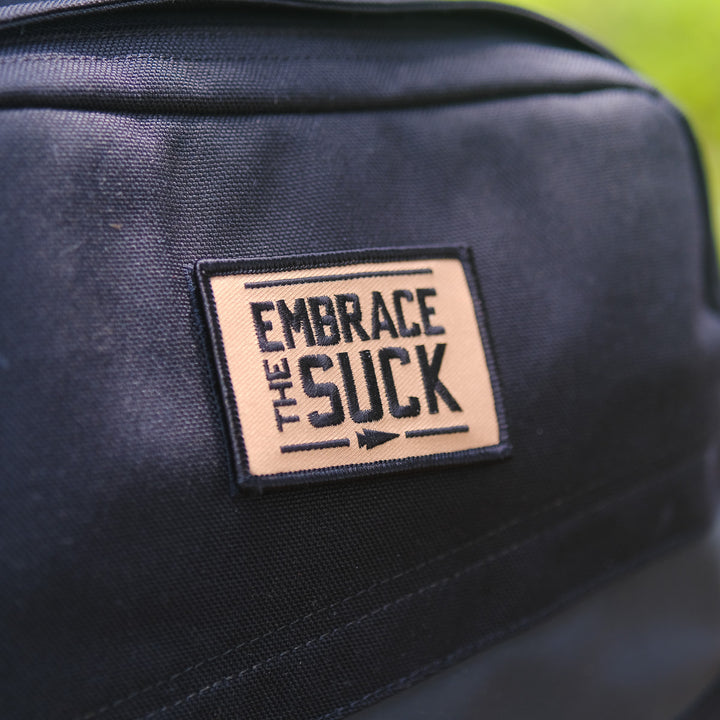 A close-up of Patch - Embrace The Suck, an embroidered patch with bold lettering and a secure VELCRO hook backing, displayed on a black backpack.