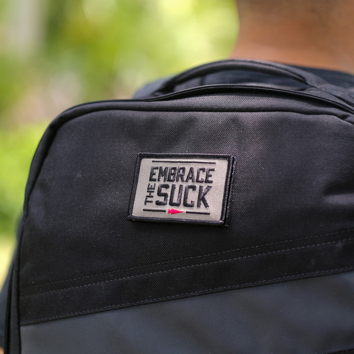 Close-up of Patch - Embrace The Suck, an embroidered patch with VELCRO hook backing for easy attachment, shown on a black backpack.