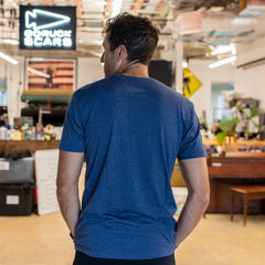 A man in a blue Embrace the Suck Tee - Tri-BlendX stands indoors facing away, with a workspace and American flag in the background—an understated nod to motivational apparel that inspires you to embrace the suck every day.