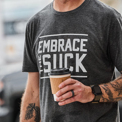 A person holds a coffee cup while wearing the Embrace the Suck Tee - Tri-Blend, featuring the USMC-inspired motto "Embrace The Suck"—a reminder to face any challenge head-on.