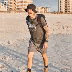 A mustachioed, tattooed man strolls the beach in a hat, backpack, and Embrace the Suck Tee - Tri-Blend—saluting a core USMC motto.