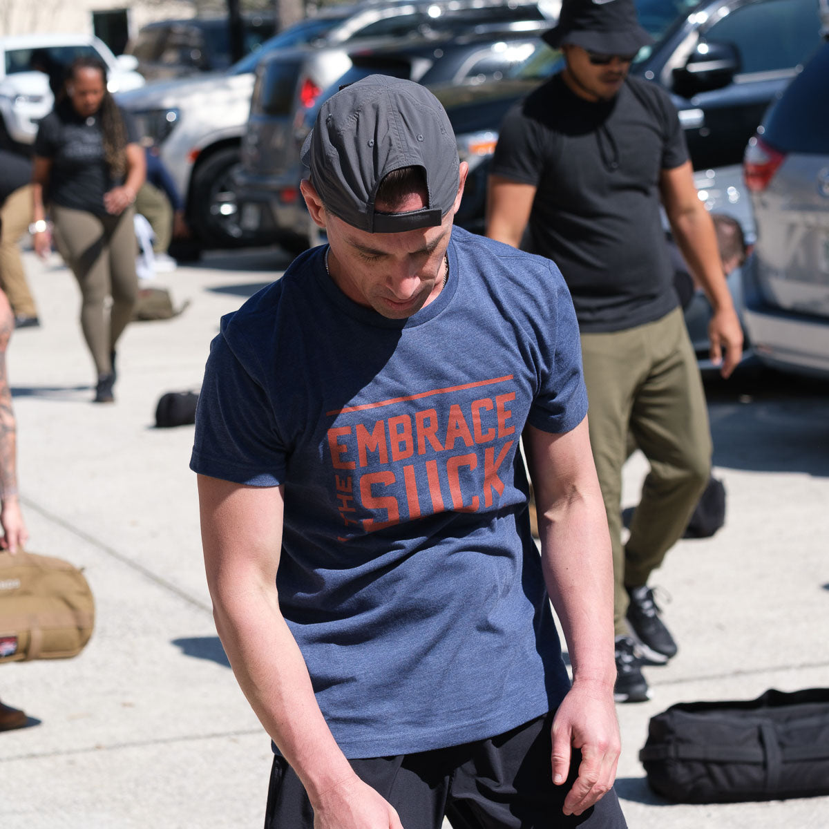 Man in a blue Embrace the Suck Tee - Tri-Blend and backwards cap stands outdoors among people and gear, sporting one of the USMC’s official mottos.