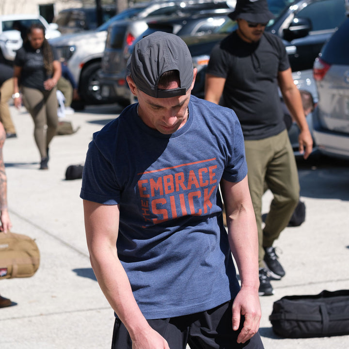 Man in a blue Embrace the Suck Tee - Tri-Blend and backwards cap stands outdoors among people and gear, sporting one of the USMC’s official mottos.