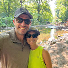 Smiling couple in sunglasses and hats poses by a sunlit creek in a leafy forest, with her wearing the Women’s USA Performance Tank made from breathable Featherweight ToughMesh fabric.