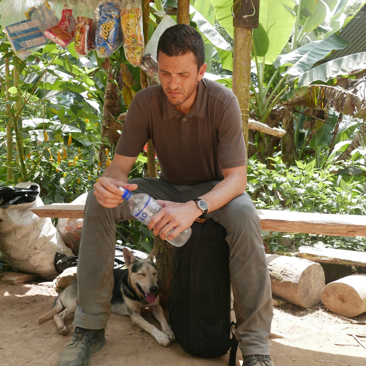 A man sits on his backpack holding a water bottle, with his dog lying beside him in a lush outdoor setting, his Spearhead Nylon Webbing Belt—USA made—providing reliable comfort for every adventure.