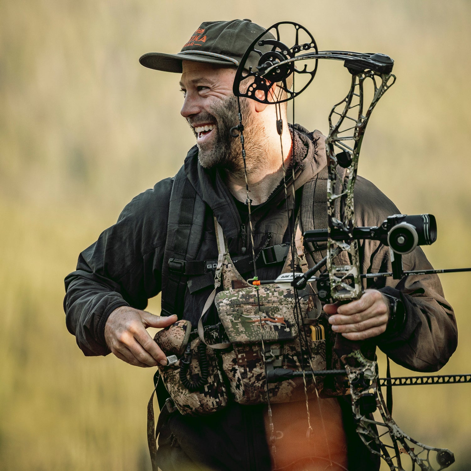 A smiling man in outdoor gear holds a camouflage compound bow in a natural setting.