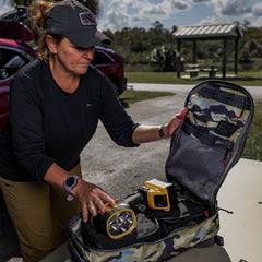 At a picnic area, a person is carefully placing equipment into the Bullet Ruck Classic - Cordura daypack by GORUCK. The padded straps promise comfort, while cars and a shelter provide the backdrop to this outdoor scene.