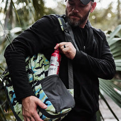 In a forest, a man wearing a black jacket and cap pulls out a red canister from the side pocket of an M24 - Cordura rucksack by GORUCK, crafted with durable materials. The scene suggests preparation or setup for an outdoor activity.
