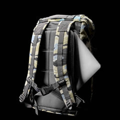 The GORUCK M24 - Cordura rucksack, showcased against a black background in a sleek camouflage pattern, reveals a laptop partially tucked into its side pocket. Constructed from robust materials, it offers padded straps and a top handle for enhanced comfort and convenience.